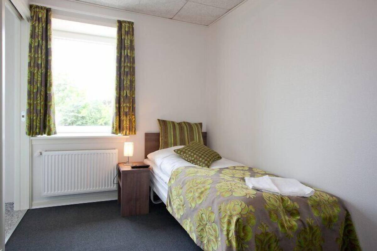 A small, neatly arranged bedroom with a single bed, a bedside table, a lamp, and a window with curtains.