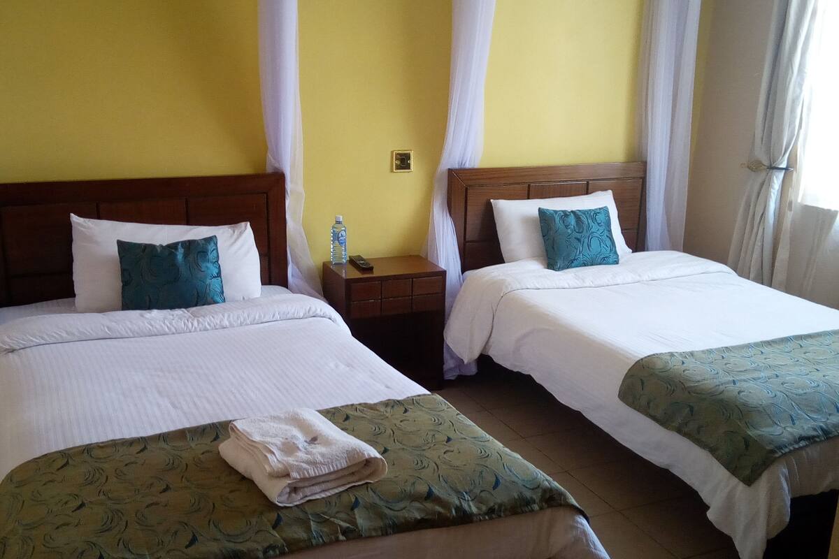 Habitación de hotel con dos camas, cada una con sábanas blancas y almohadas color aguamarina. Se observa un mesita de noche con una botella de agua y un control remoto, además de una toalla doblada sobre la cama.