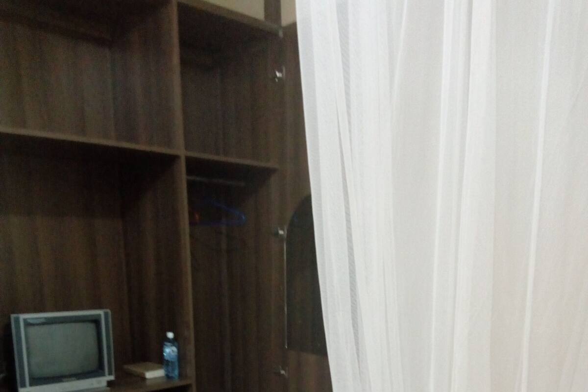 Un dormitorio con un armario de madera, una cama con ropa de cama blanca, un televisor sobre una estantería y una cortina entreabierta.