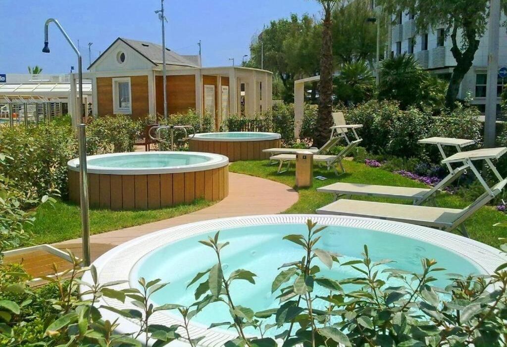 A pool area with two hot tubs, lounge chairs, and a wooden building with a sloped roof.