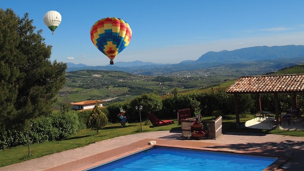 Zona de piscina con una pérgola y mobiliario exterior, con vistas a un paisaje pintoresco que incluye montañas y un globo aerostático a lo lejos.