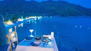 Ein Tisch ist für das Abendessen gedeckt, mit Blick auf einen Strand und Berge im Dämmerlicht.