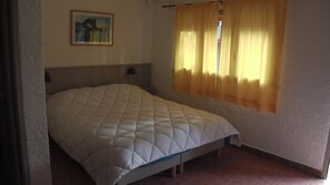 A single bed with a quilted mattress in a room with a plain wall and a window with yellow curtains. - village vacances KALLISTERRA (Zonza)