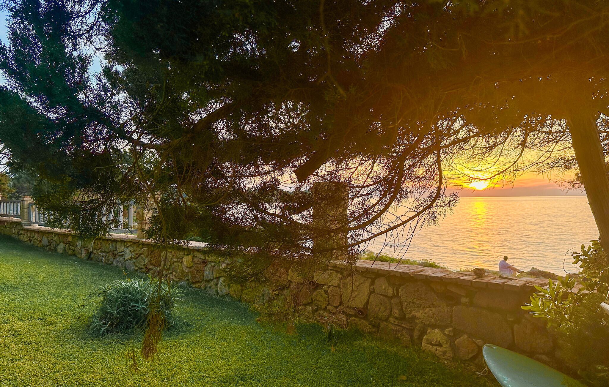 A stone wall with a grassy area and trees in the foreground, overlooking a body of water at sunset.