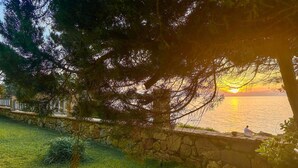 A stone wall with a grassy area and trees in the foreground, overlooking a body of water at sunset. - Beach front apartment in Leventoxori, Ilias (Leventoxori, Ilias)
