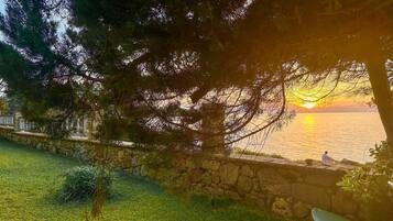 A stone wall with a grassy area and trees in the foreground, overlooking a body of water at sunset.