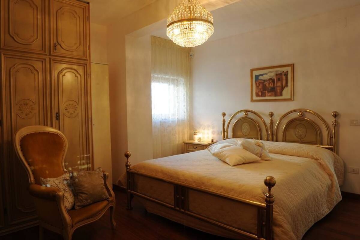 Quarto com uma cama grande, uma cadeira de madeira, um candelabro e um quadro na parede.