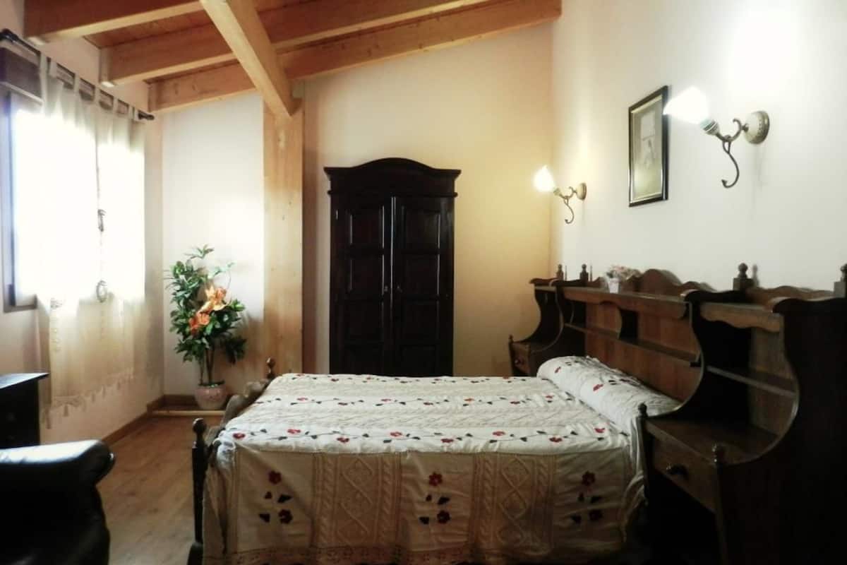 Une chambre avec un plafond en bois, un lit recouvert d’une couvre-lit à motifs floraux, une armoire en bois foncé, une table de chevet avec une lampe et une plante en pot.