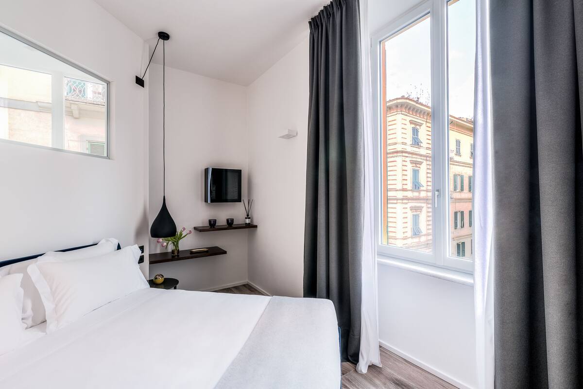 Una moderna camera d'albergo con un letto grande, una TV su un mobile, una finestra con tende e vista sugli edifici circostanti.