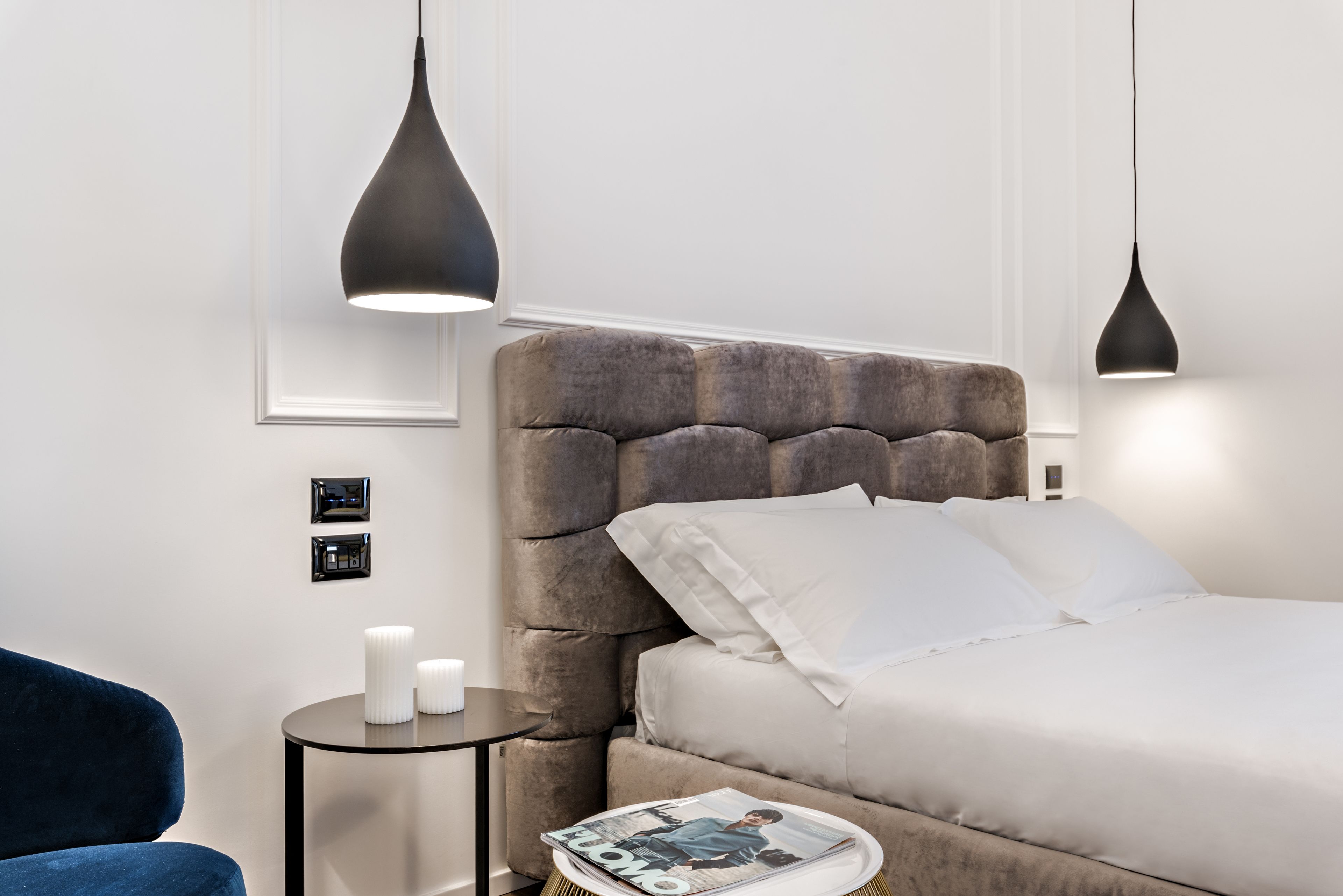 Una camera da letto moderna con una testiera trapuntata, un tavolino rotondo con una rivista e due lampade a sospensione.