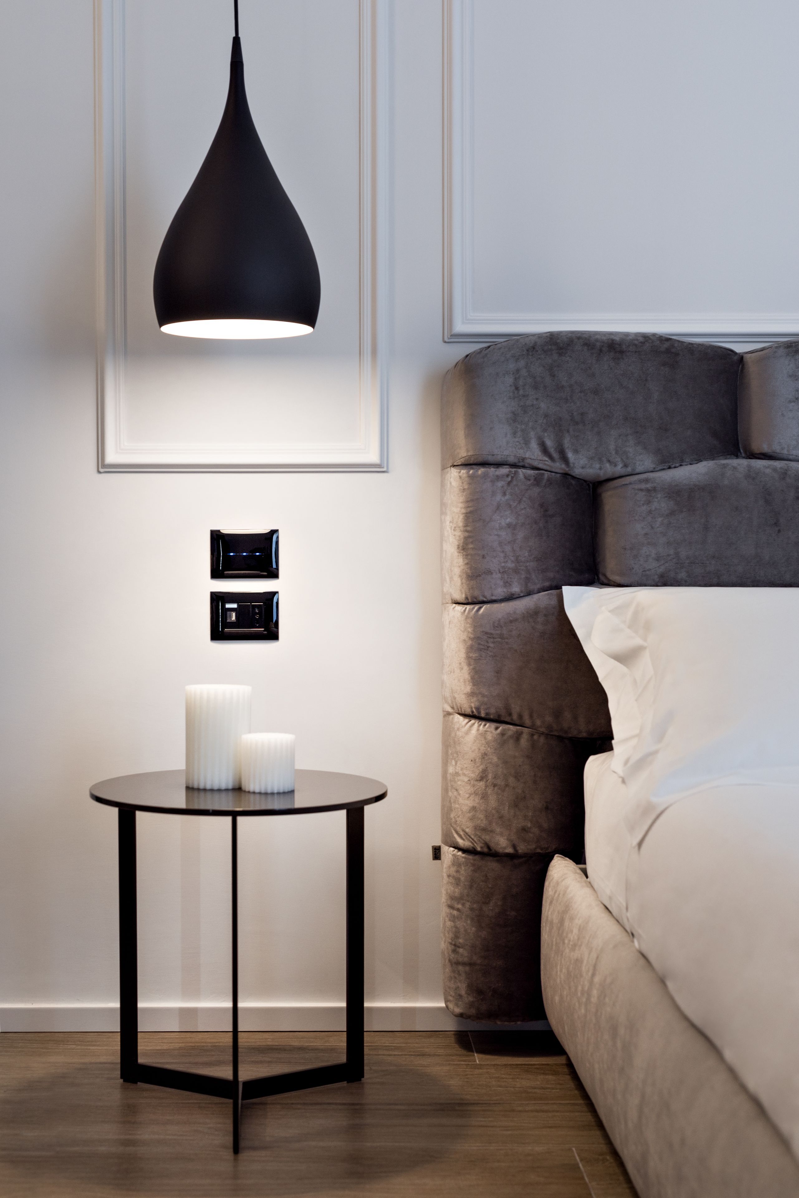 A modern bedroom with a tufted headboard, a round black side table with a white vase, and a pendant light.