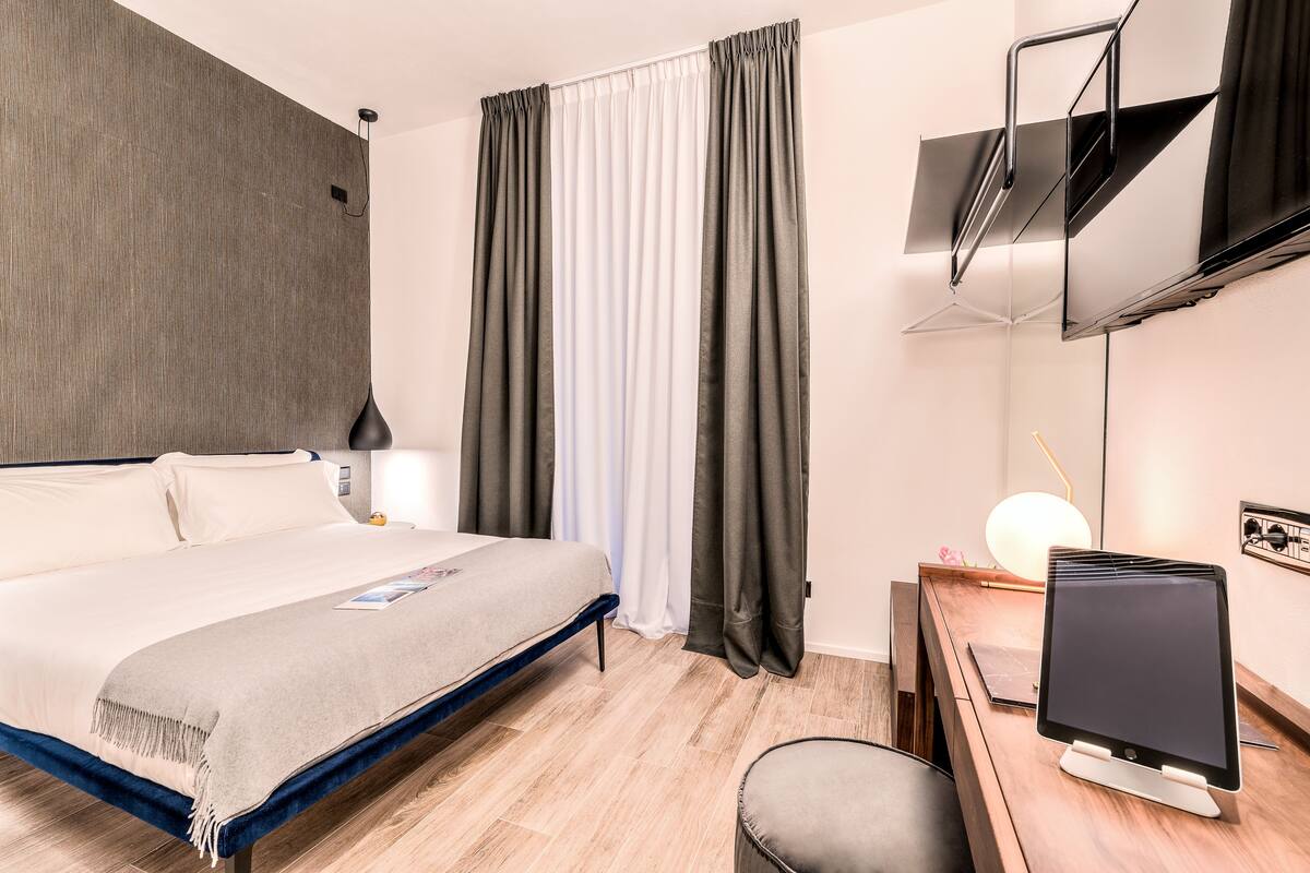 Una camera d'albergo moderna con un letto, una scrivania, una TV a schermo piatto e un'ampia finestra con tende.