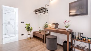 A modern room with a wooden desk, a grey ottoman, a wall-mounted TV, and a small shelf with glasses.