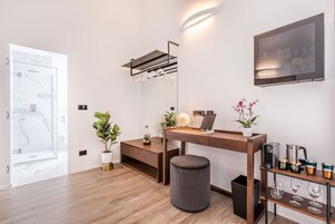 Una habitación moderna con un escritorio de madera, un reposapiés gris, un televisor montado en la pared y una pequeña estantería con vasos.