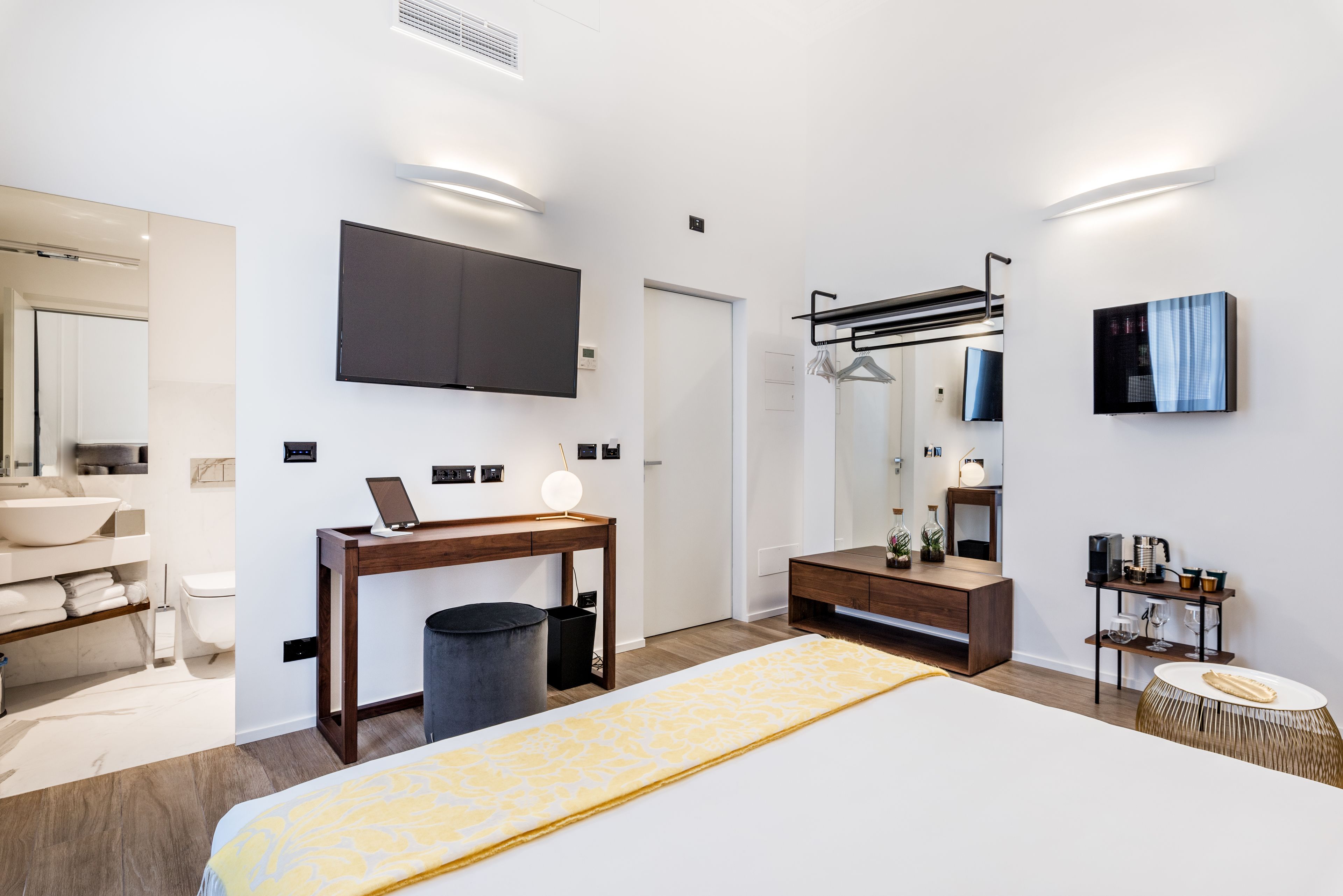 A modern hotel room with a large bed, a desk with a laptop, a TV mounted on the wall, a vanity with a mirror, and a small side table with a lamp.