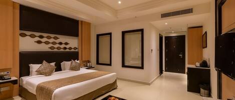 A modern hotel room with a large bed, a bedside table with a fruit bowl, a desk with a chair, and a TV on the wall.