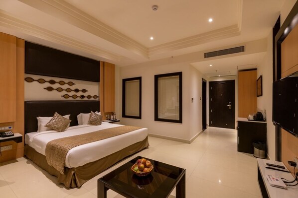 A modern hotel room with a large bed, a bedside table with a fruit bowl, a desk with a chair, and a TV on the wall. - Cygnett Lite (Alwar)