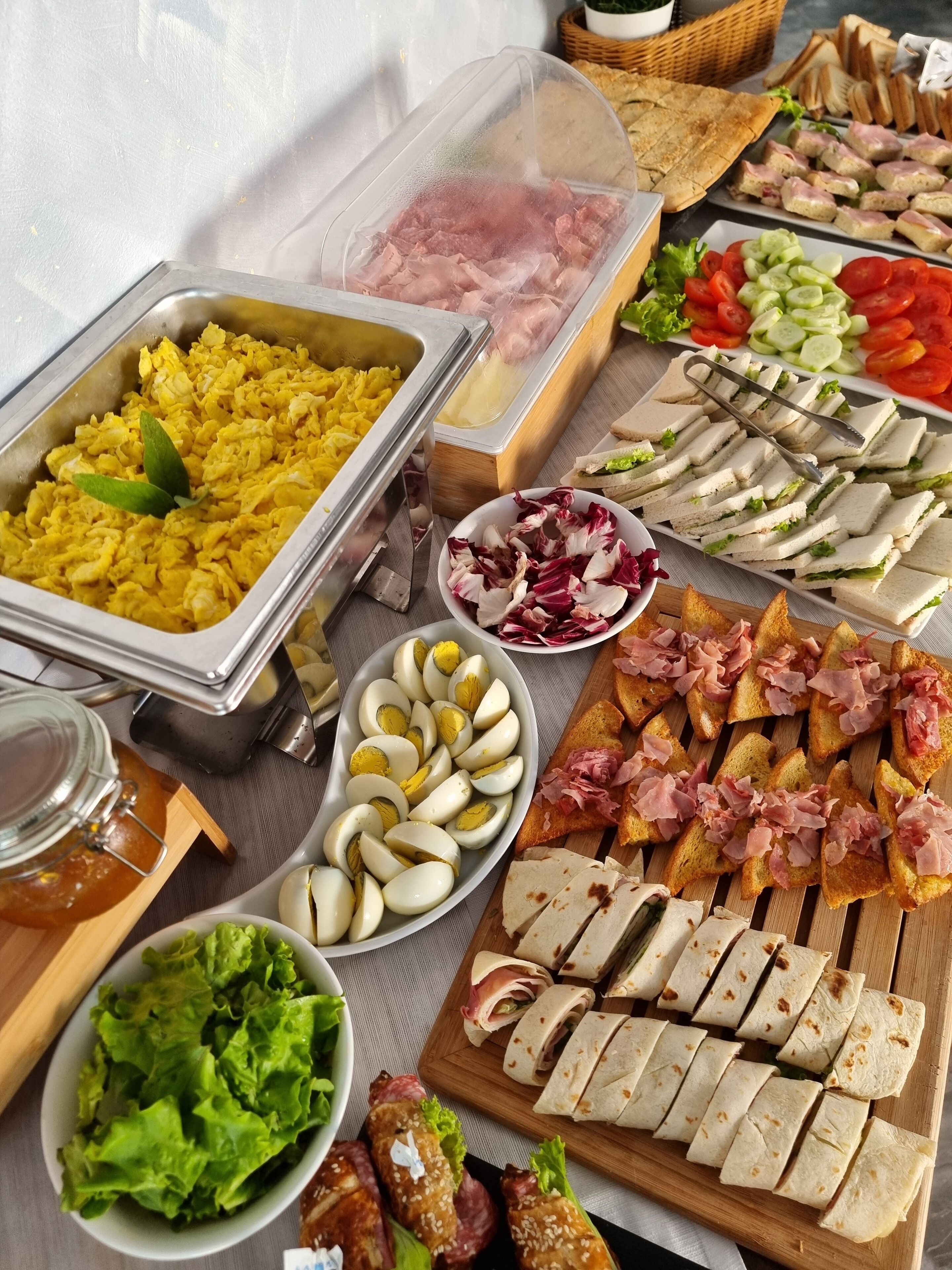 Un bufé con diversos alimentos, incluyendo huevos revueltos, carnes frías, sándwiches y verduras.