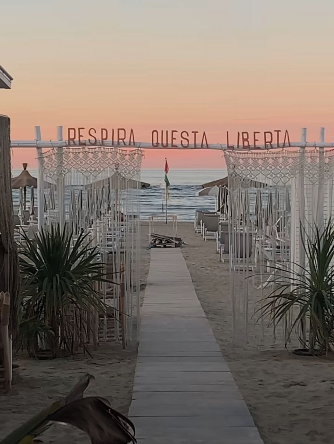 A beachside walkway leading to a seating area with a sign reading "RESPIRA QUESTA LIBERTA".