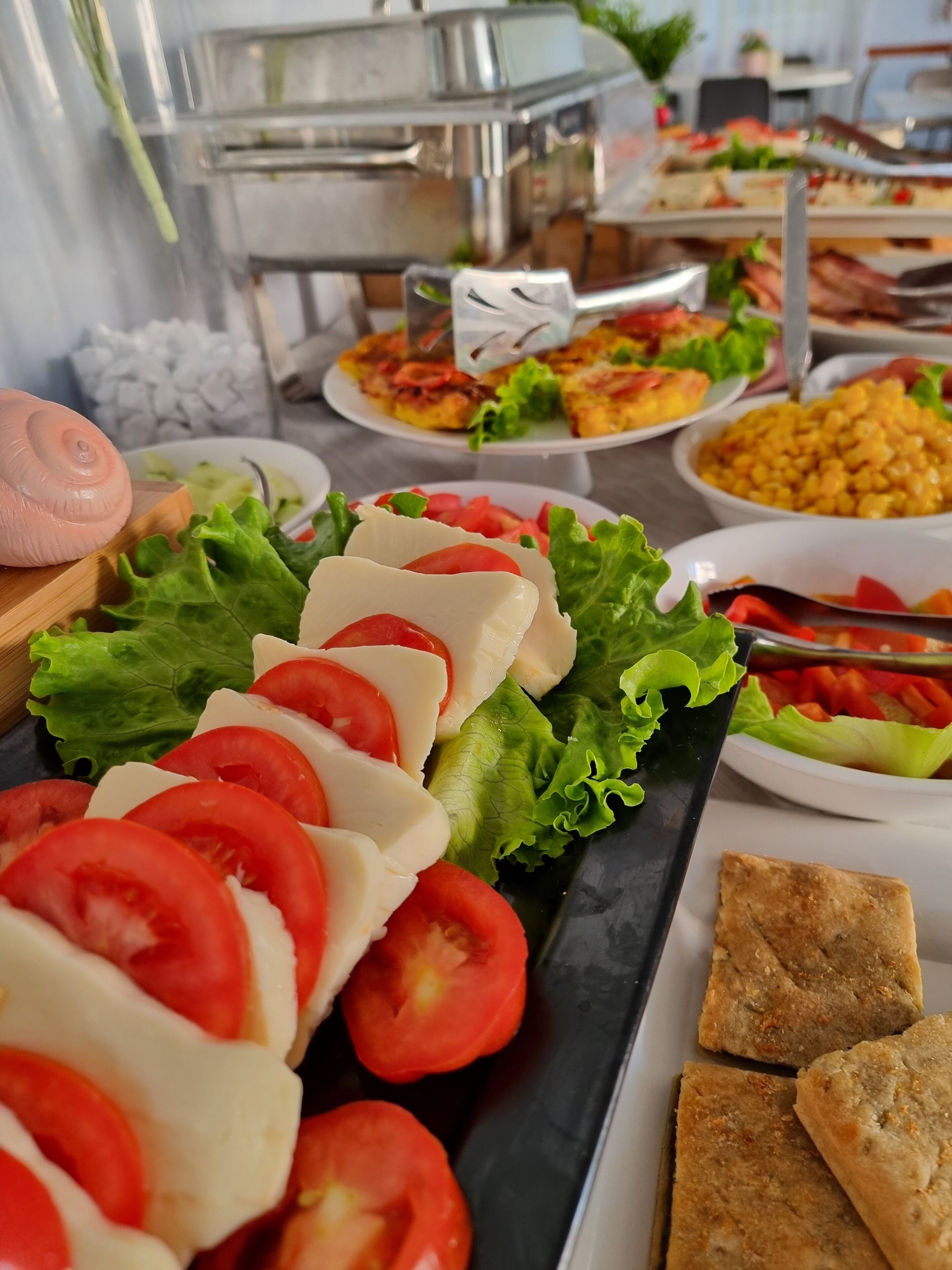 A buffet with various food items including sliced tomatoes, cheese, lettuce, and corn.