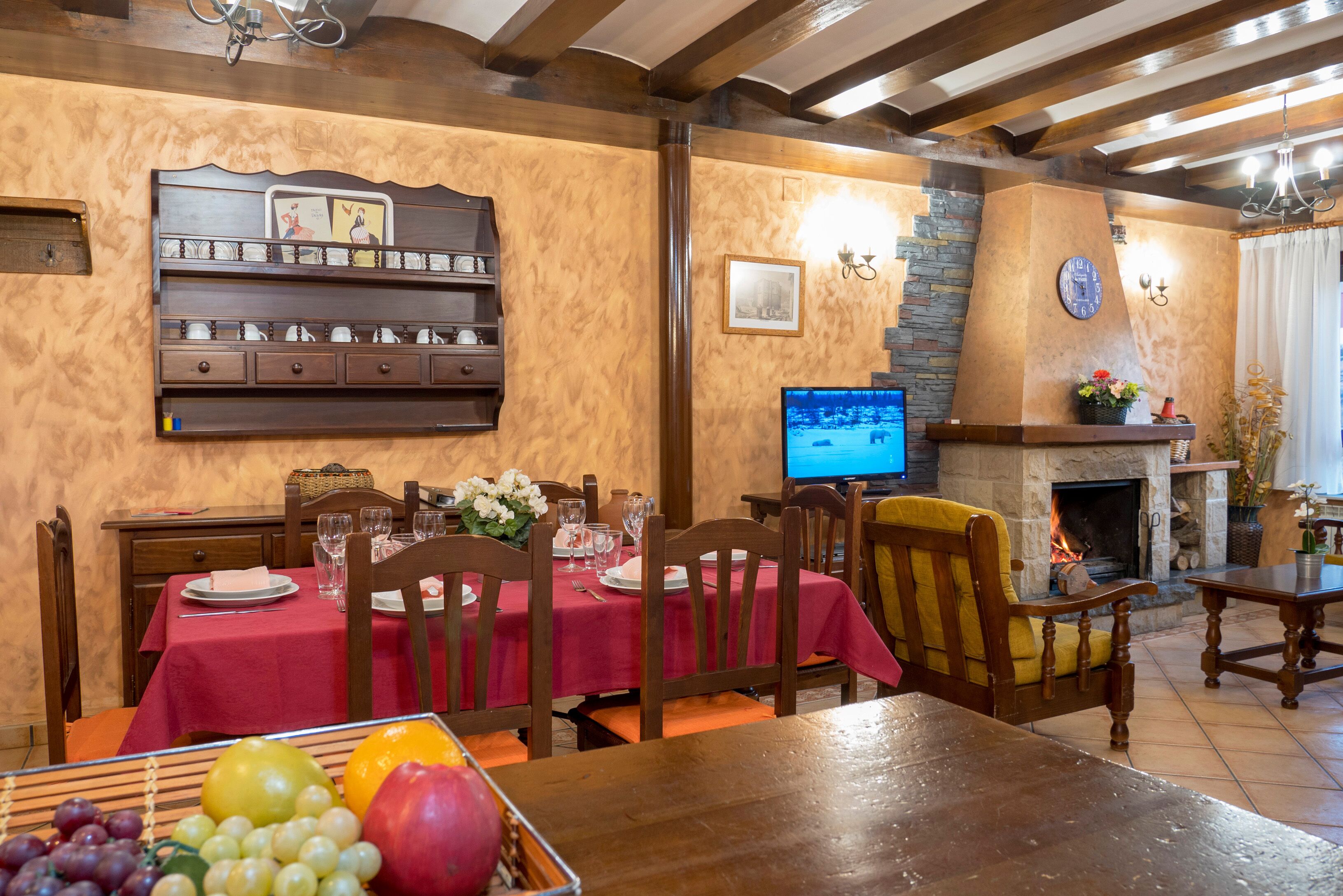 A dining area with a red tablecloth, wooden chairs, a fruit basket, and a fireplace.