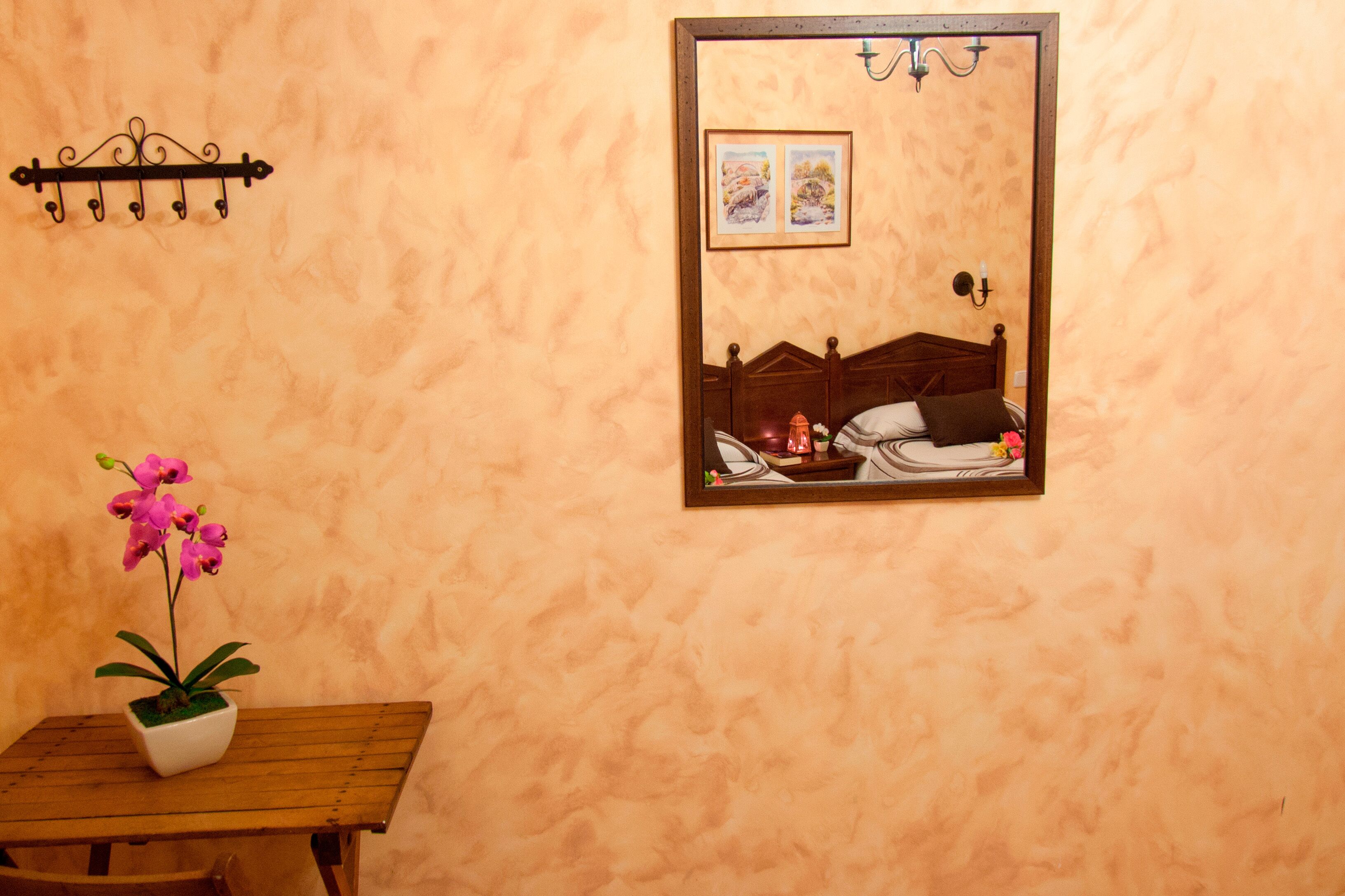 Une pièce avec un lit en bois, une petite table sur laquelle est posée une orchidée en pot, un miroir fixé au mur qui reflète le lit et un tableau, et un luminaire fixé au mur.
