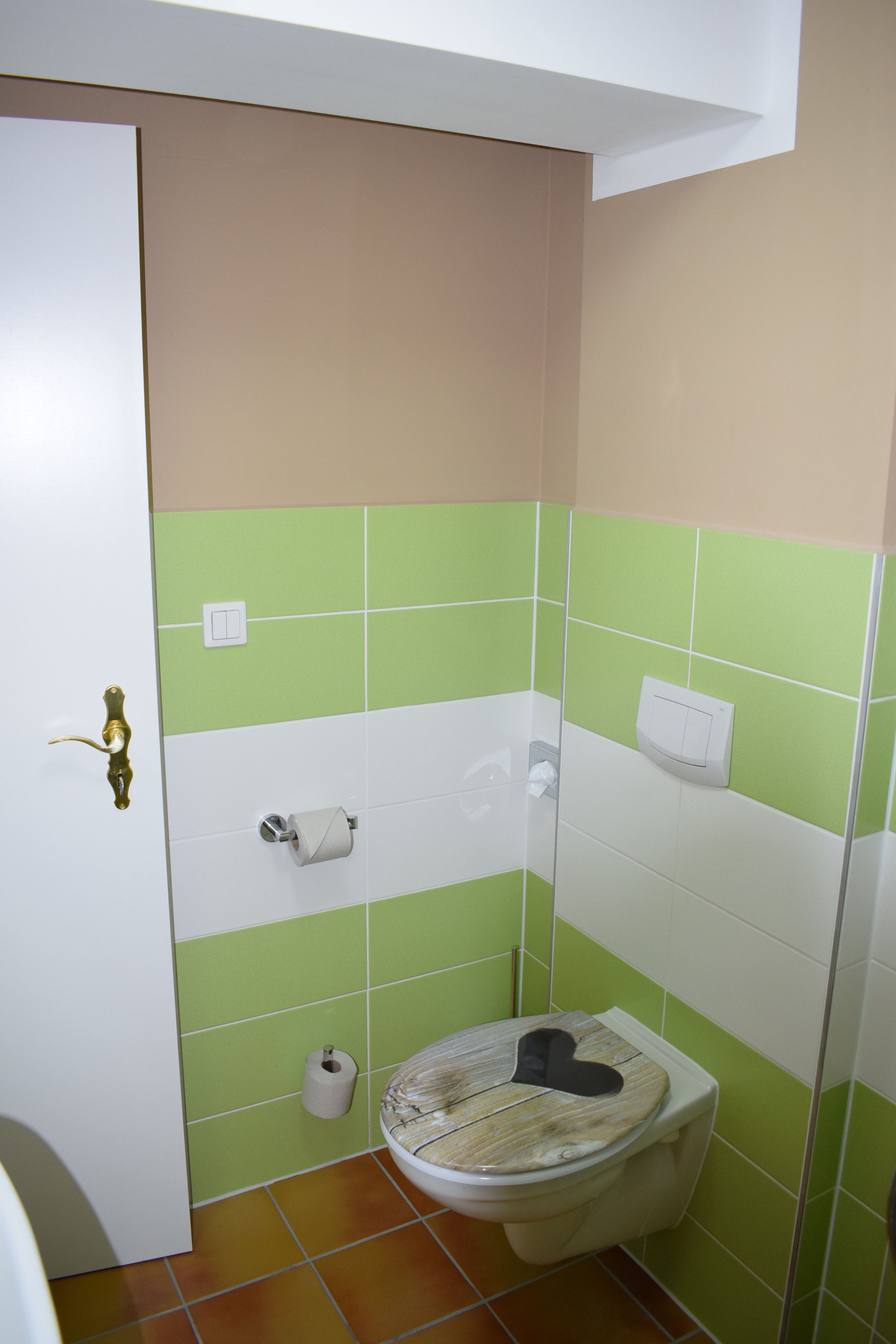 A bathroom with green and beige tiles, a toilet with a paper towel holder, and a door with a brass handle.