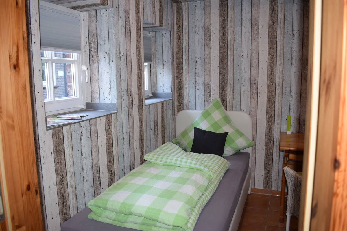 Una cama individual con colcha verde a cuadros y almohada negra, una mesita de noche de madera con espejo, y una ventana con vista a un edificio de ladrillo.