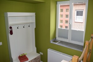 Quarto com parede verde, radiador branco, janela com persianas, armário branco com ganchos e toalha vermelha no balcão.
