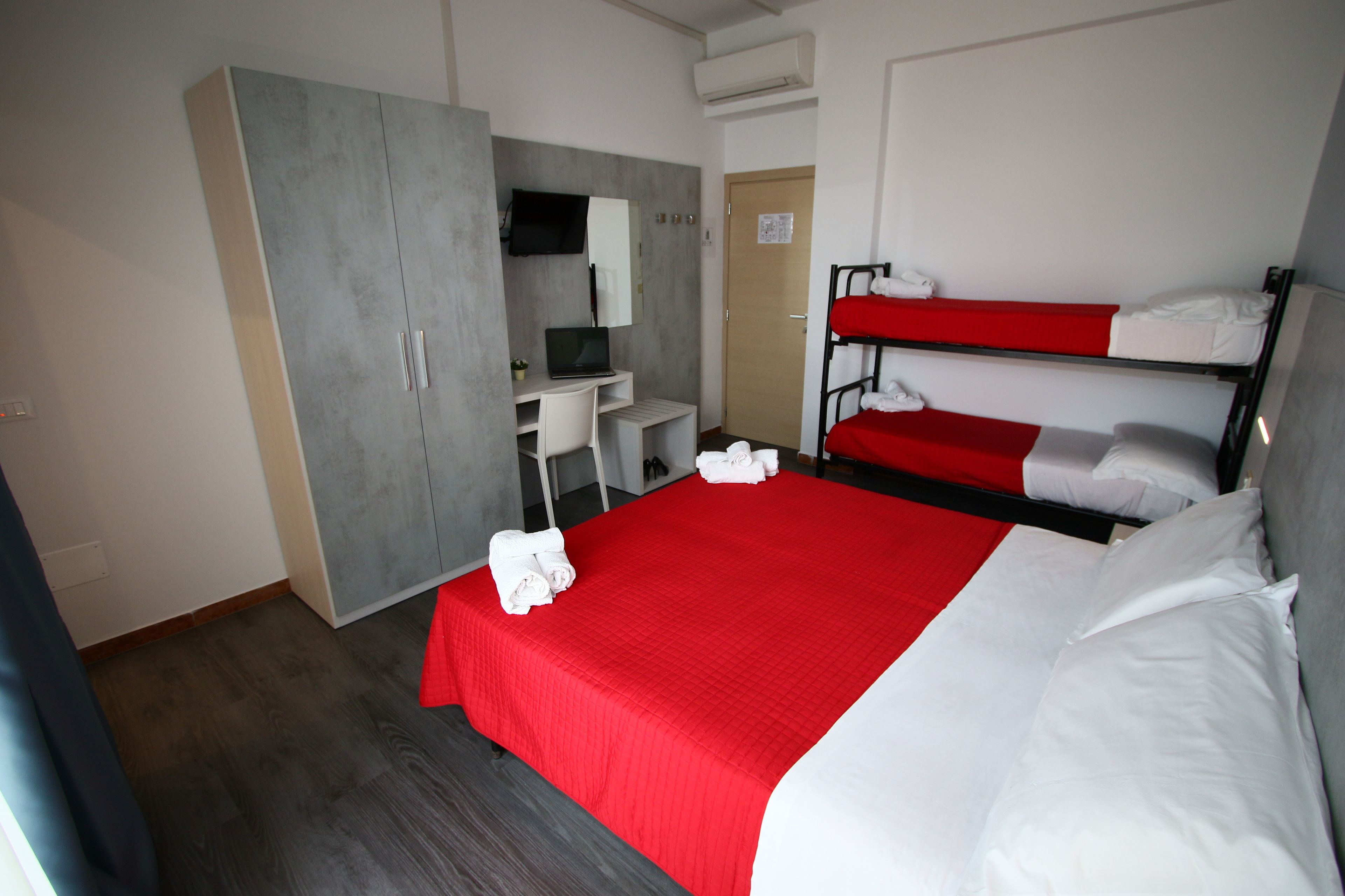 Quarto de hotel com uma cama beliche, uma mesa com cadeira, um guarda-roupa e uma televisão na parede.