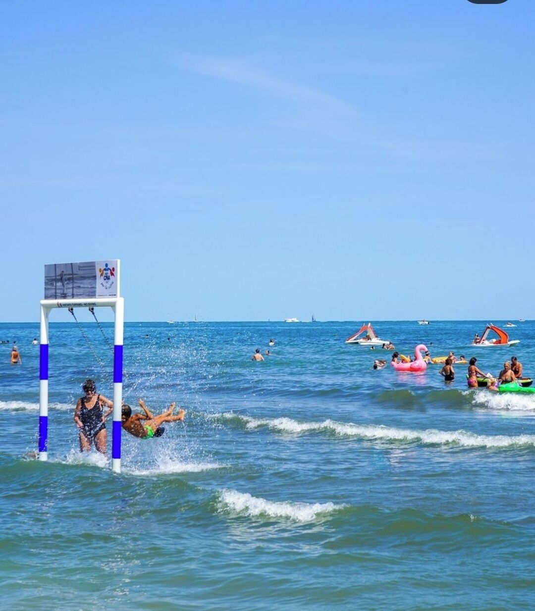 Eine Strandansicht mit einem Tor, Personen, die schwimmen und treiben, und Booten im Hintergrund.