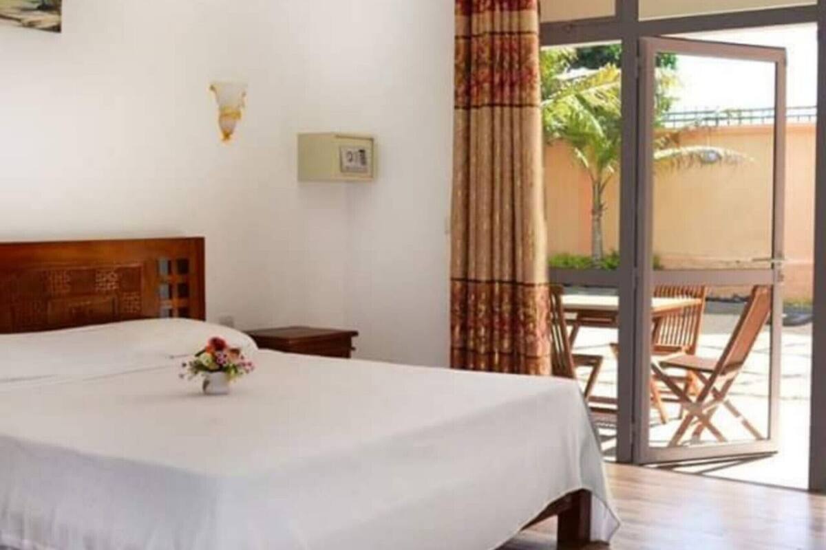 A hotel room with a large bed, a small table with a vase of flowers, a wall-mounted light, and a view of the outside through a glass door.