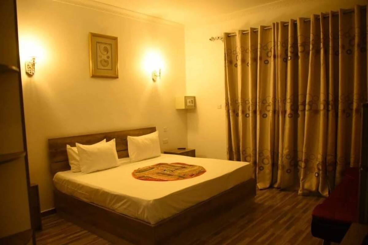 A hotel room with a bed, a nightstand, a wall-mounted lamp, a framed picture, and a window with curtains.