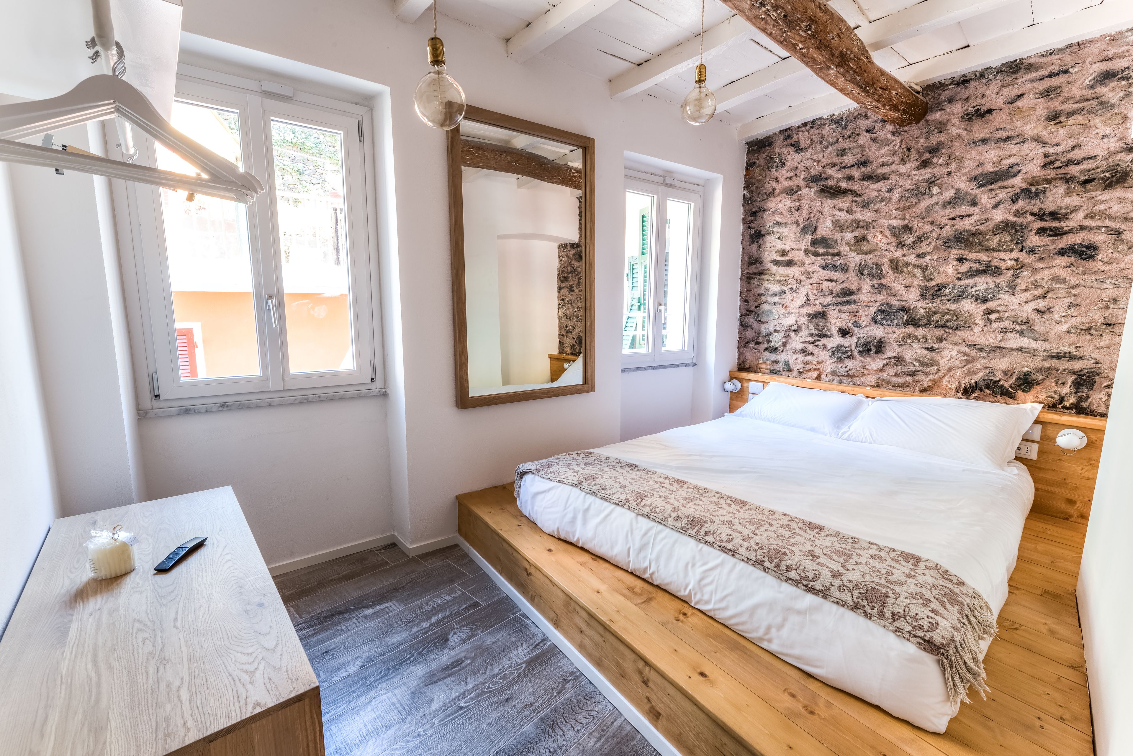 A bedroom with a bed, a wooden bench, a bedside table, a mirror, and a stone wall.