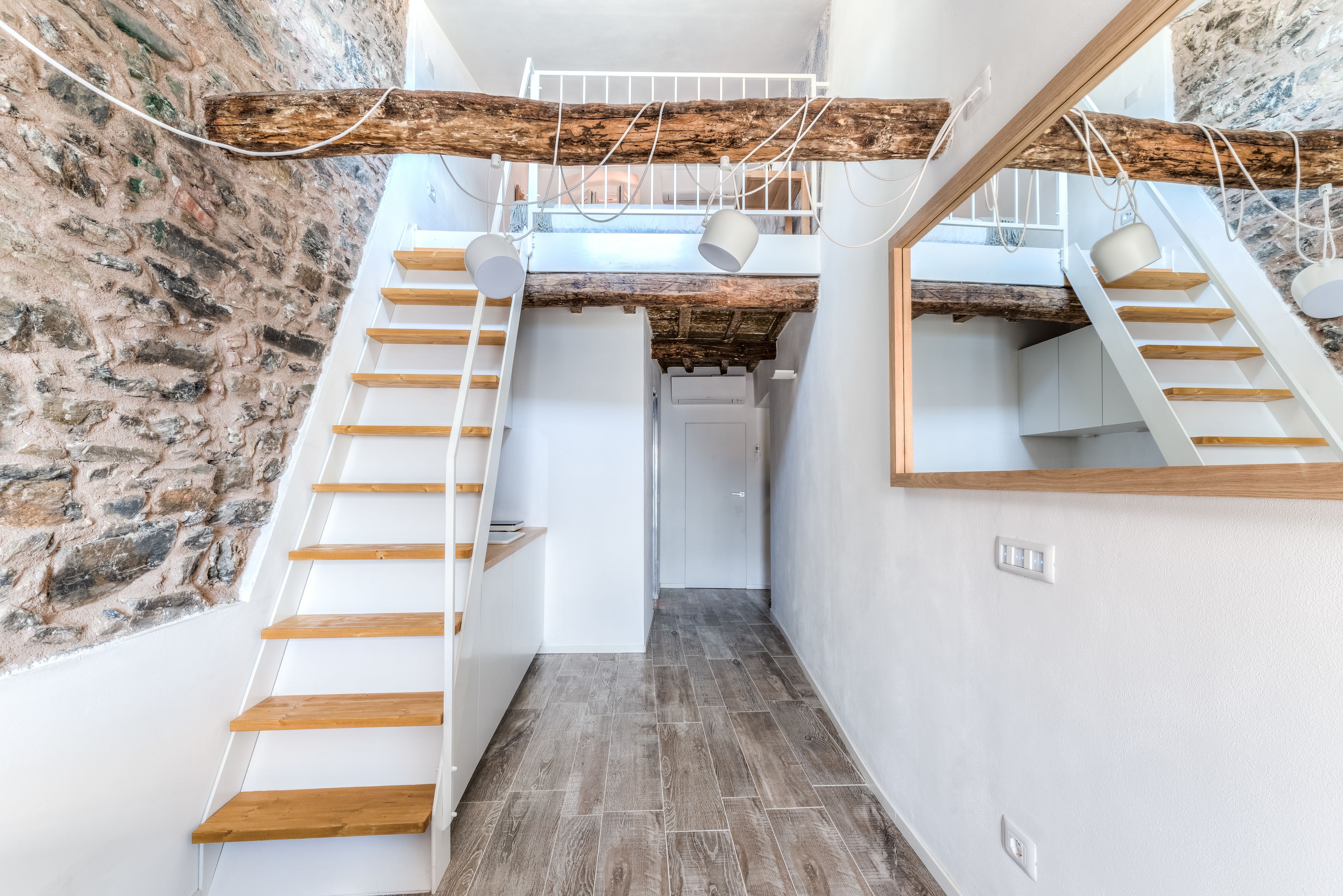 A modern interior with a wooden staircase, a stone wall, and a loft area with a wooden beam ceiling.