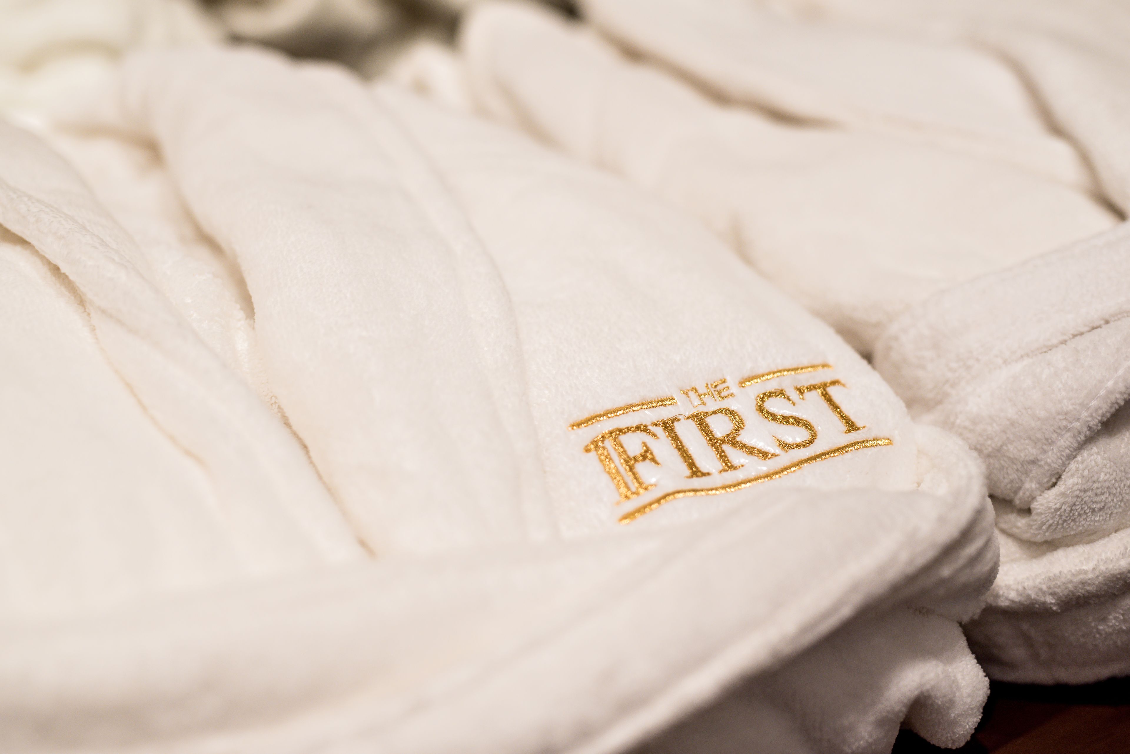 Una pila de toallas blancas con un logotipo dorado bordado que dice "THE FIRST".