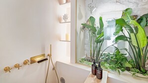 A modern bathroom with a bathtub, a glass shower enclosure, and a large mirror reflecting a view of plants and a window.