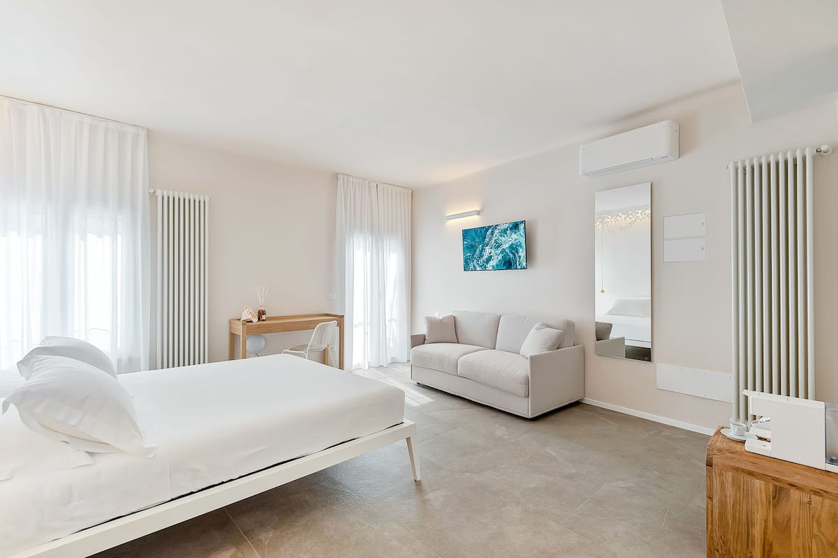 A modern hotel room with a bed, a sofa, a desk, and a large window with curtains.