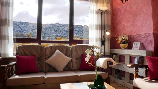A cozy living room with a sofa, a table, and a bookshelf. A window offers a view of snow-covered hills. - Casa Rural Los Moros (Solosancho)