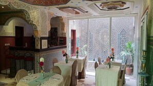 A dining area with ornate ceiling and walls, tables set with white tablecloths and floral centerpieces, and a view through a patterned screen. - Hotel Cabourg (Dakar)