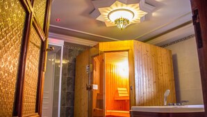A sauna room with a wooden door and bench, a shower area with a glass door, and a ceiling light fixture. - Hotel Cabourg (Dakar)
