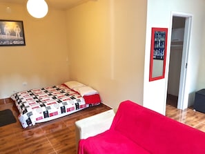 A small, well-lit room with a bed, a red sofa, and a door leading to another room. - Suite Capivari Room (Campos do Jordão)