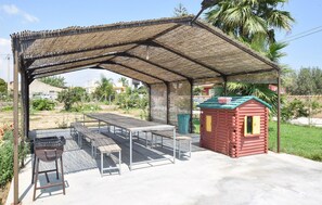 Relax in a serene outdoor space with a covered seating area, long table, and comfortable chairs, surrounded by lush vegetation and a charming playhouse.