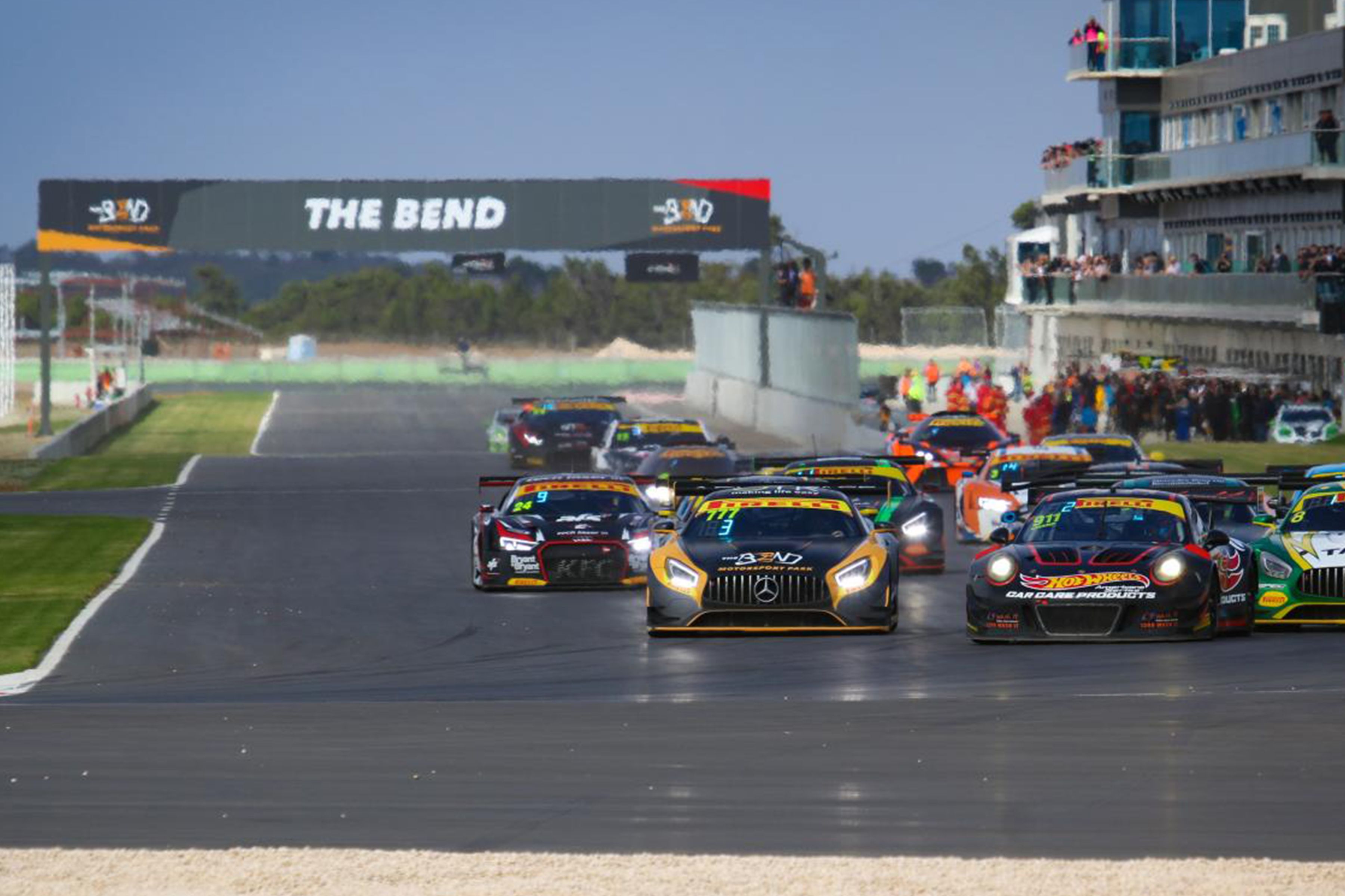 A racing event with multiple cars on a track, spectators in the background, and a large banner reading "THE BEND".