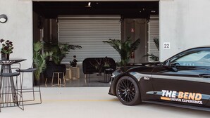 A black sports car parked outside a building with a garage door, number 22, and a sign reading "THE BEND DRIVER EXPERIENCE". - Rydges Pit Lane Hotel (Tailem Bend)