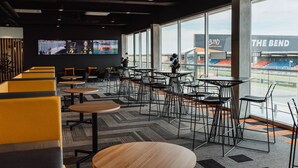 A modern indoor seating area with tables and chairs, large windows, and a view of an outdoor stadium. - Rydges Pit Lane Hotel (Tailem Bend)