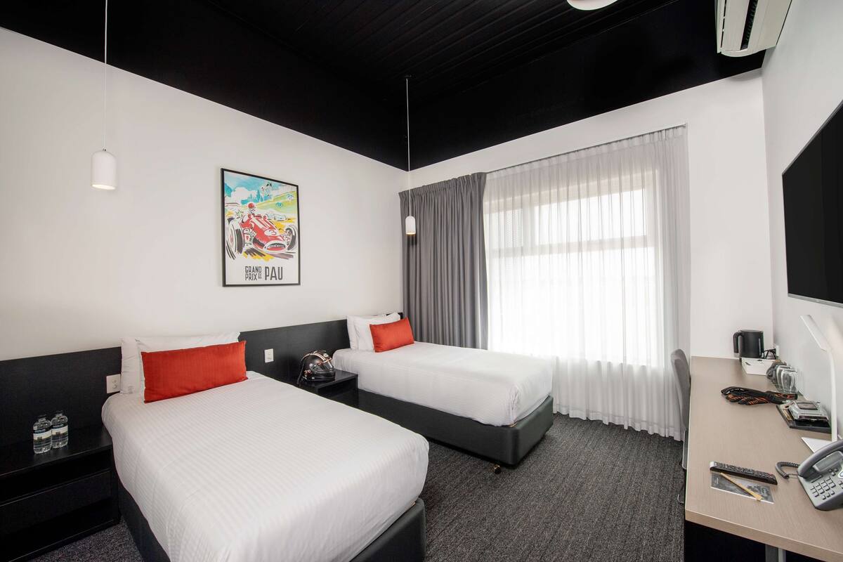 A hotel room with two beds, a desk with a telephone, a chair, a window with curtains, and a poster on the wall.