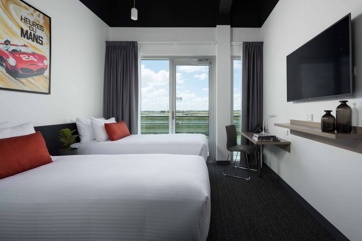 A hotel room with two beds, a TV, a desk with a chair, and a view of the outdoors.