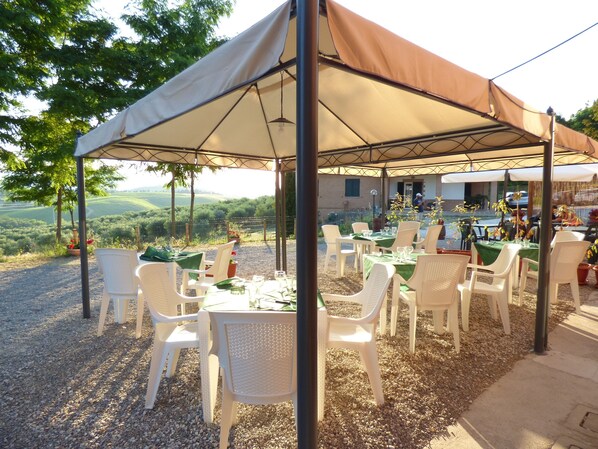 An outdoor dining area with white plastic chairs and tables under a large canopy, set on a gravel surface with a view of rolling hills in the background. - Agriturismo Il Sambuco (Monteriggioni)