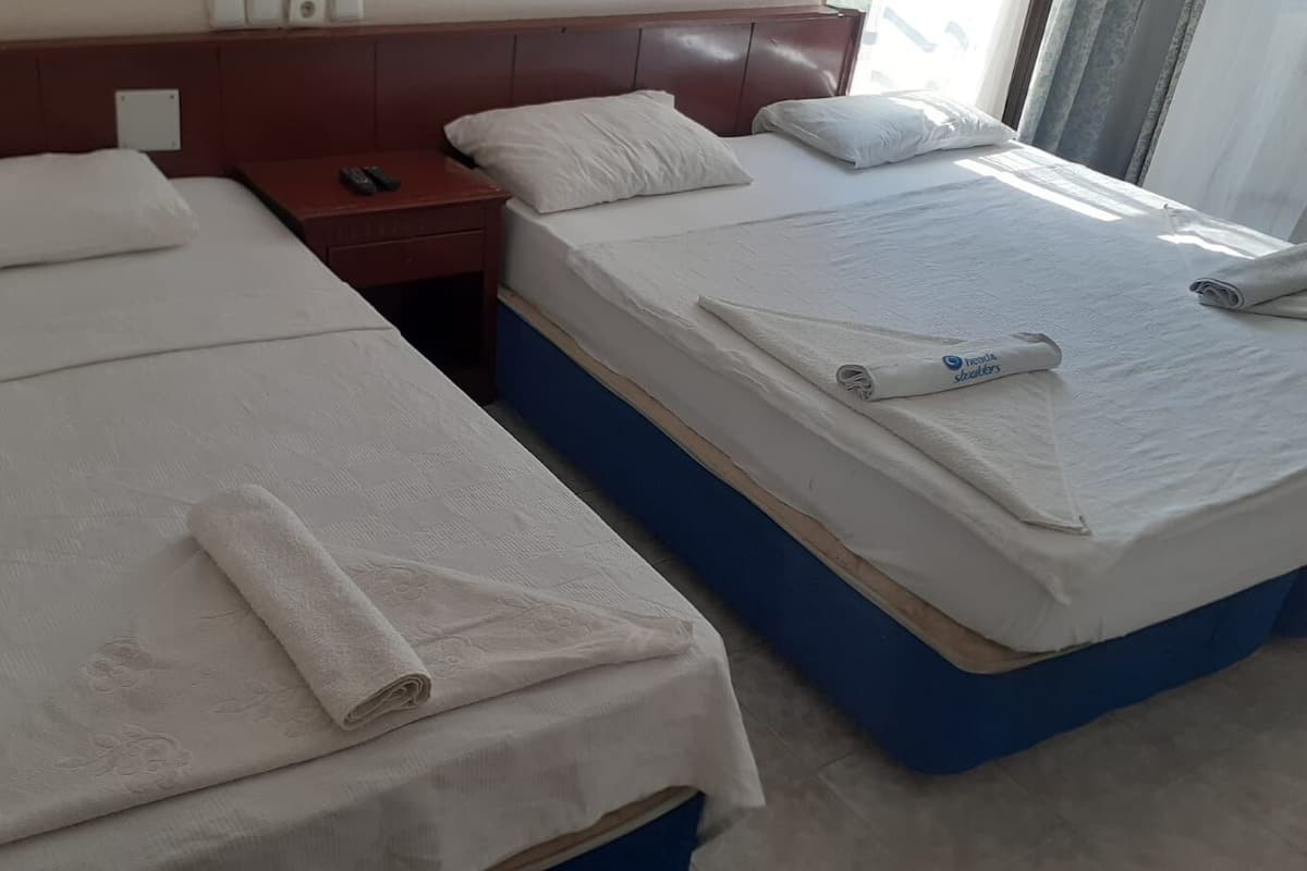 Dos camas individuales con ropa blanca y toallas, un cabecero de madera, una mesita de noche con control remoto y una ventana con cortinas.